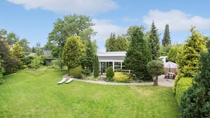 Une maison moderne dotée d’une grande façade vitrée, entourée d’un jardin bien entretenu et de grands arbres.