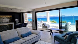 Ein modernes Wohnzimmer mit einer großen Couch, einer kleineren Couch und einem Couchtisch. Es gibt eine Küchenzeile mit Steinrückwand und einen Essbereich mit weißen Stühlen. Das Zimmer verfügt über große Fenster mit Blick auf das Meer und die Berge.