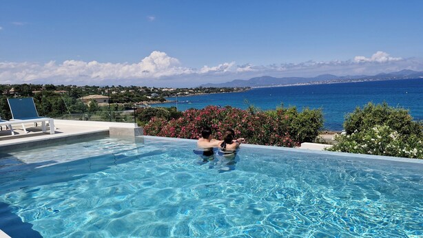 Ein klares, blaues Schwimmbecken mit zwei Personen im Wasser, umgeben von viel Grün und Blick auf das Meer sowie entfernte Berge.