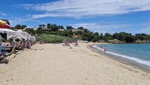 Ein sandiger Strand mit Sonnenschirmen und Liegestühlen, ein klarer Himmel und eine felsige Küste mit Bäumen.