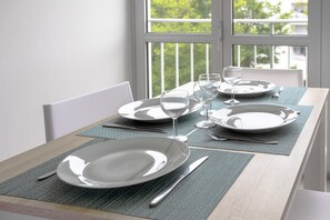 Mesa de comedor dispuesta con platos, vasos y cubiertos, cerca de una ventana con vista a follaje.