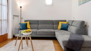 Una sala de estar con un sofá gris, cojines amarillos y azules, una mesa de centro de vidrio con un cuenco de fruta, una lámpara de pie y un radiador.