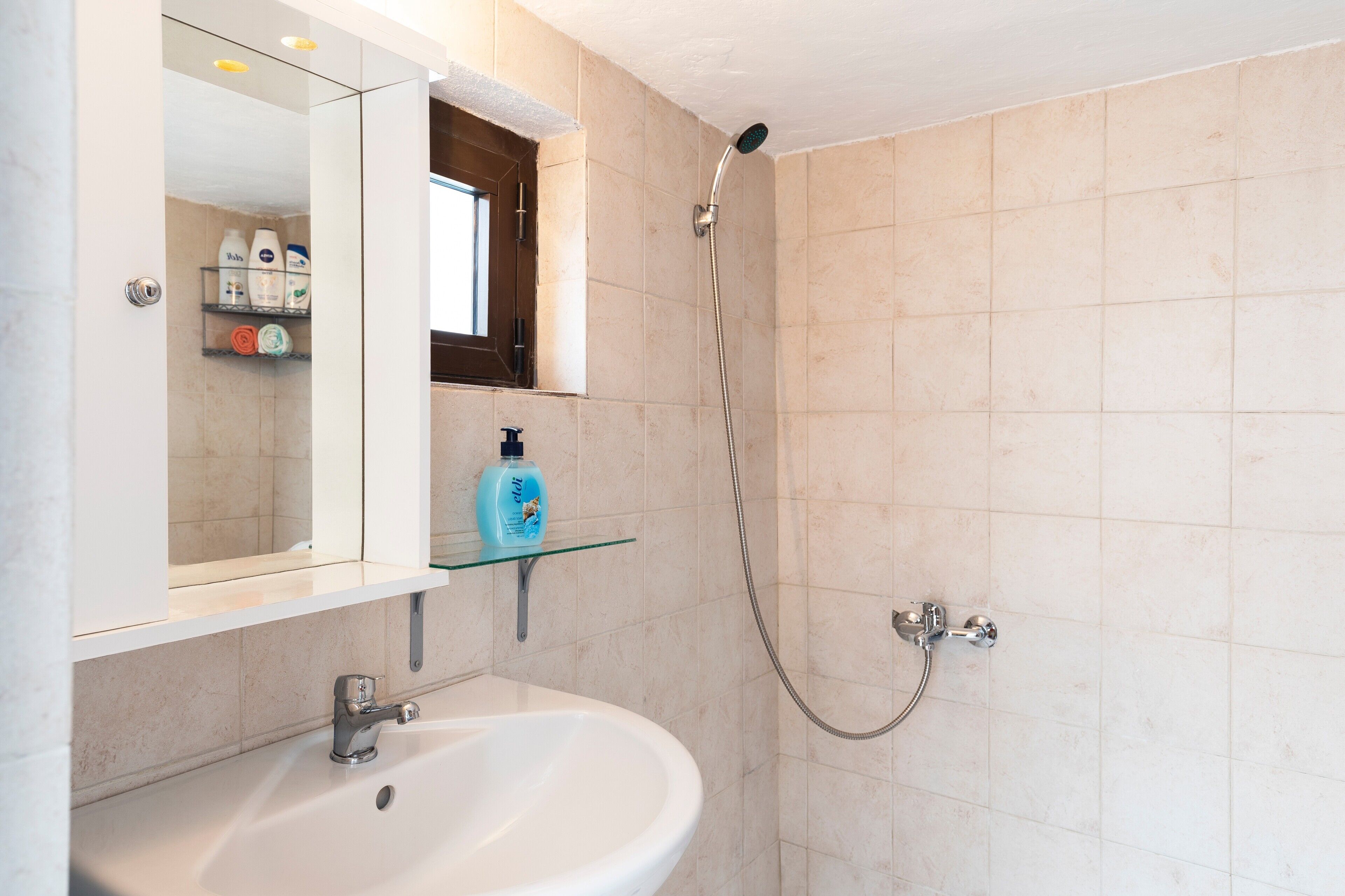 Un baño con lavamanos blanco, un estante de vidrio con artículos de aseo, ducha con cabezal de ducha manual y un espejo con ventana arriba.