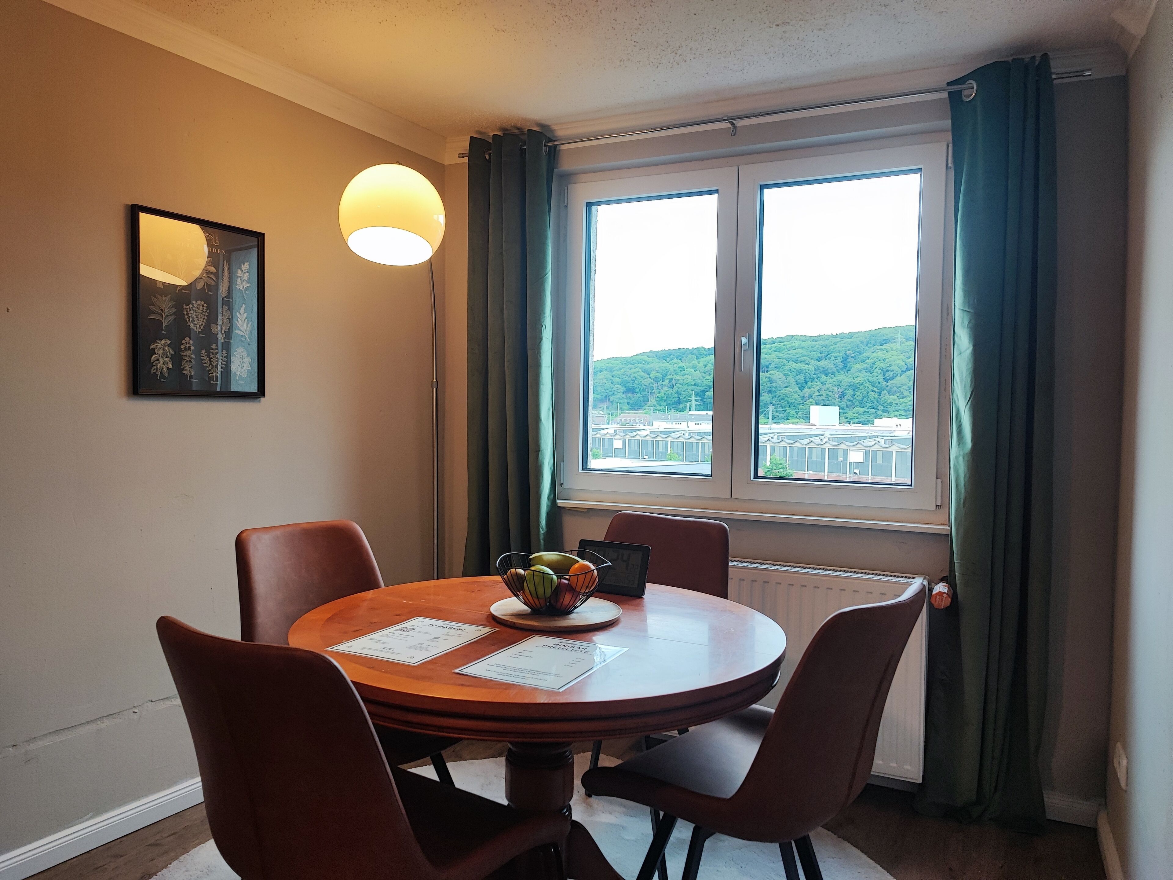 Un coin repas avec une table ronde en bois, quatre chaises, une estampe botanique encadrée au mur, un lampadaire et une fenêtre avec des rideaux.