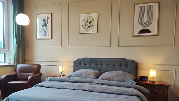 Un dormitorio ordenado con una cama grande, dos mesitas de noche con lámparas, una silla y cuadros en la pared.