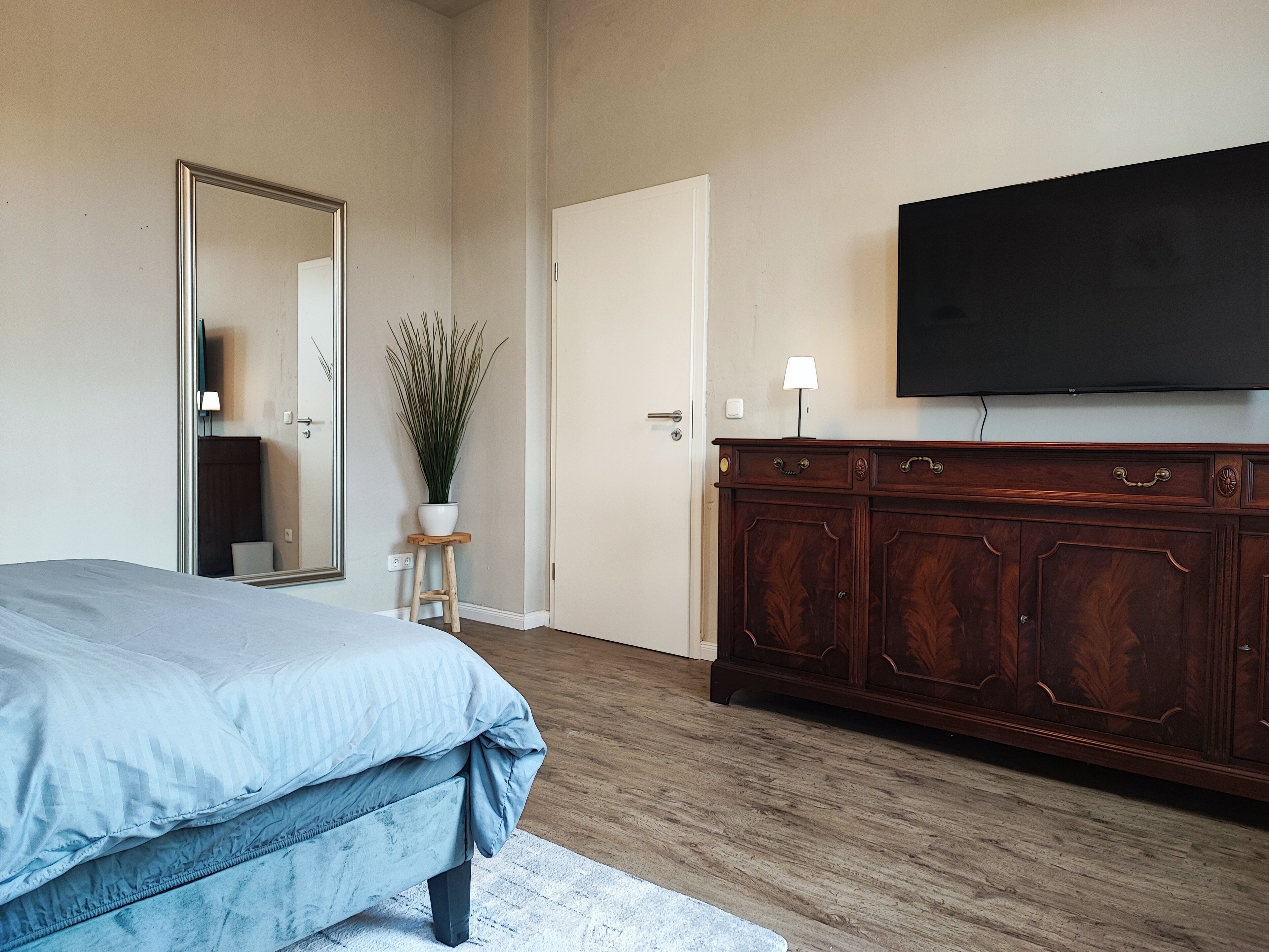 A bedroom with a bed, a dresser, a mirror, a potted plant, and a TV on the wall.