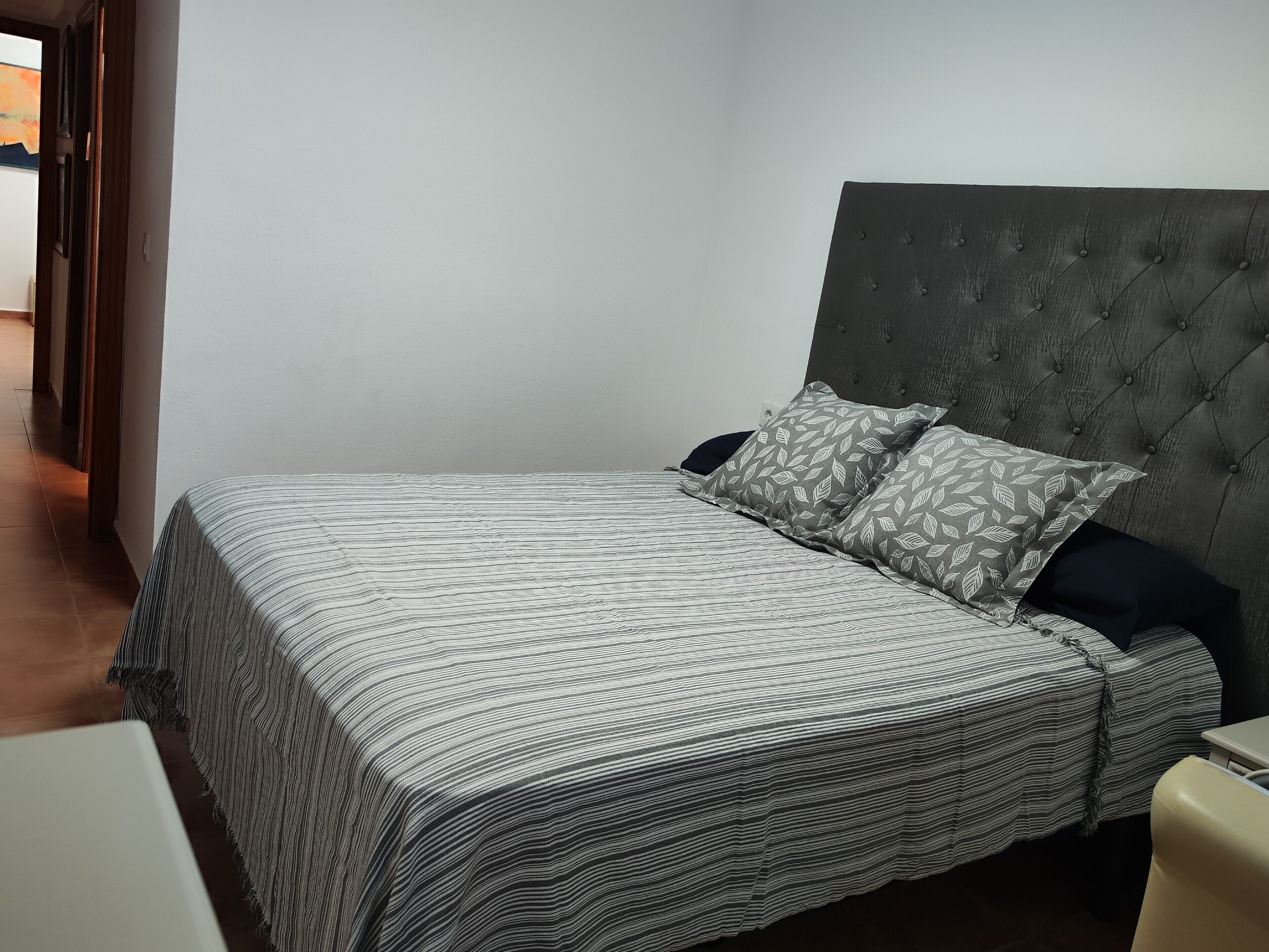 A neatly made bed with a striped bedspread and two patterned pillows, a tufted headboard, and a plain wall.