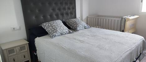 A bedroom with a tufted headboard, two pillows with leaf patterns, a white knitted bedspread, a white nightstand with drawers, and a radiator.