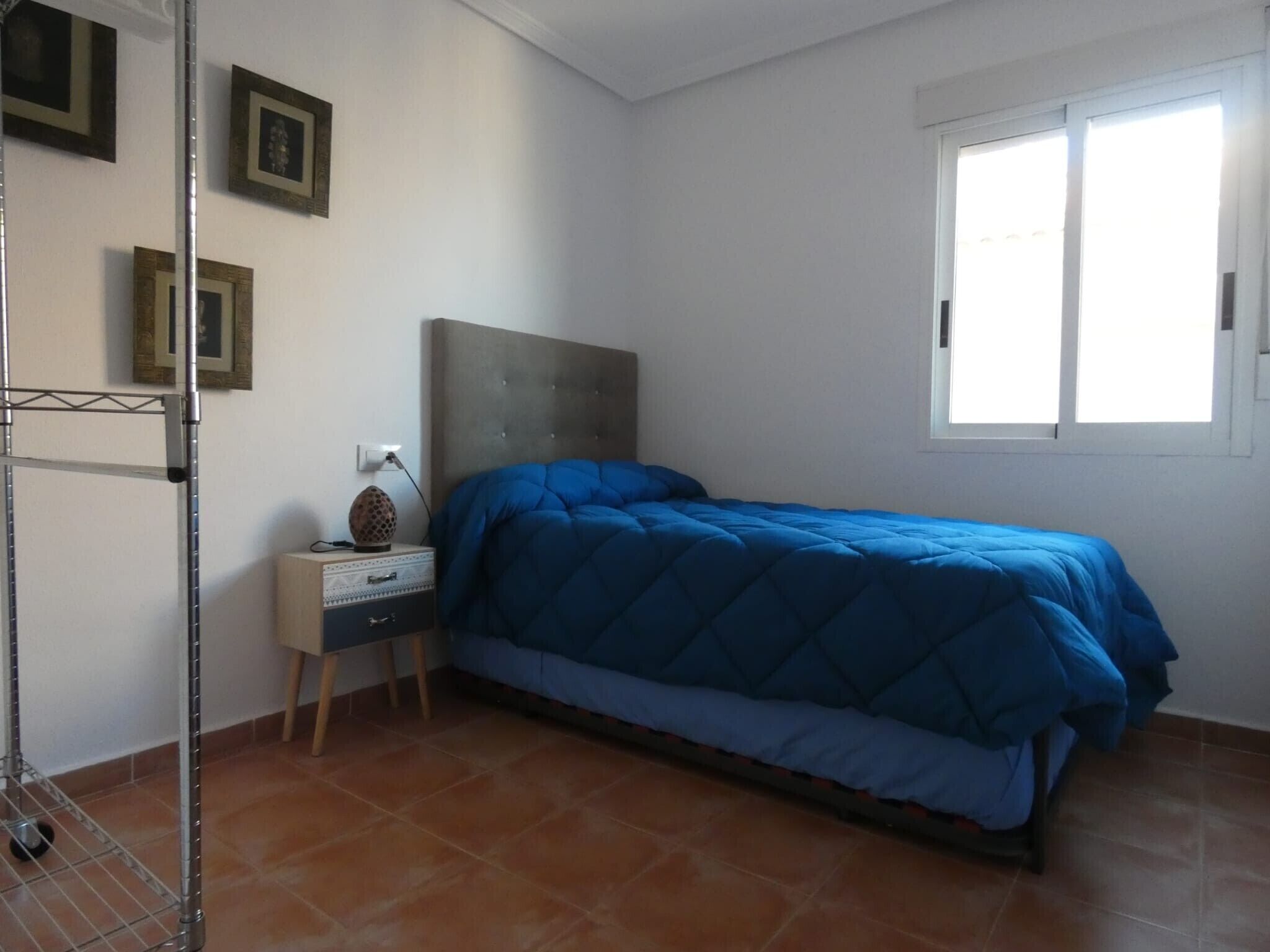 A bedroom with a bed, a nightstand, a wall-mounted shelf, and a window.