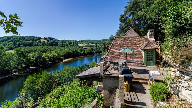 Une maison de campagne avec une cheminée en pierre, une porte verte et une terrasse dotée de meubles d’extérieur.