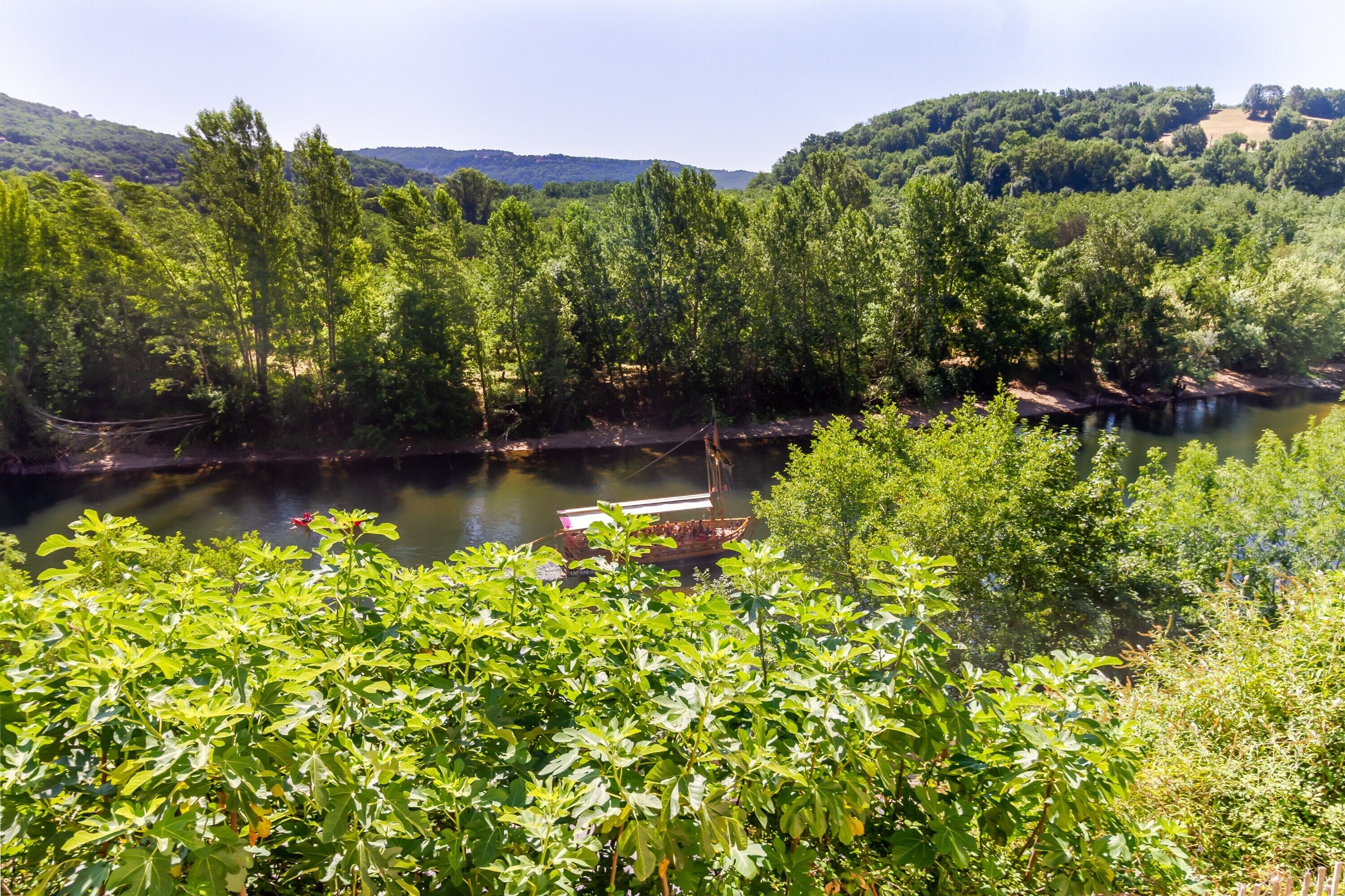 A river with boats, surrounded by dense greenery and hills in the background.