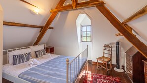 Une chambre avec un plafond en pente, un lit recouvert d’une courtepointe à motifs bleus et blancs, une chaise en bois, un radiateur et une petite fenêtre.