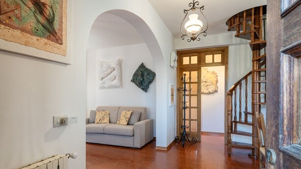 Un pasillo con radiador, un sofá gris con cojines estampados, una escalera de madera y una puerta con marco decorativo.