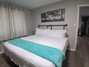 Una camera da letto con un letto, tende bianche, un quadro raffigurante alberi e un corridoio.