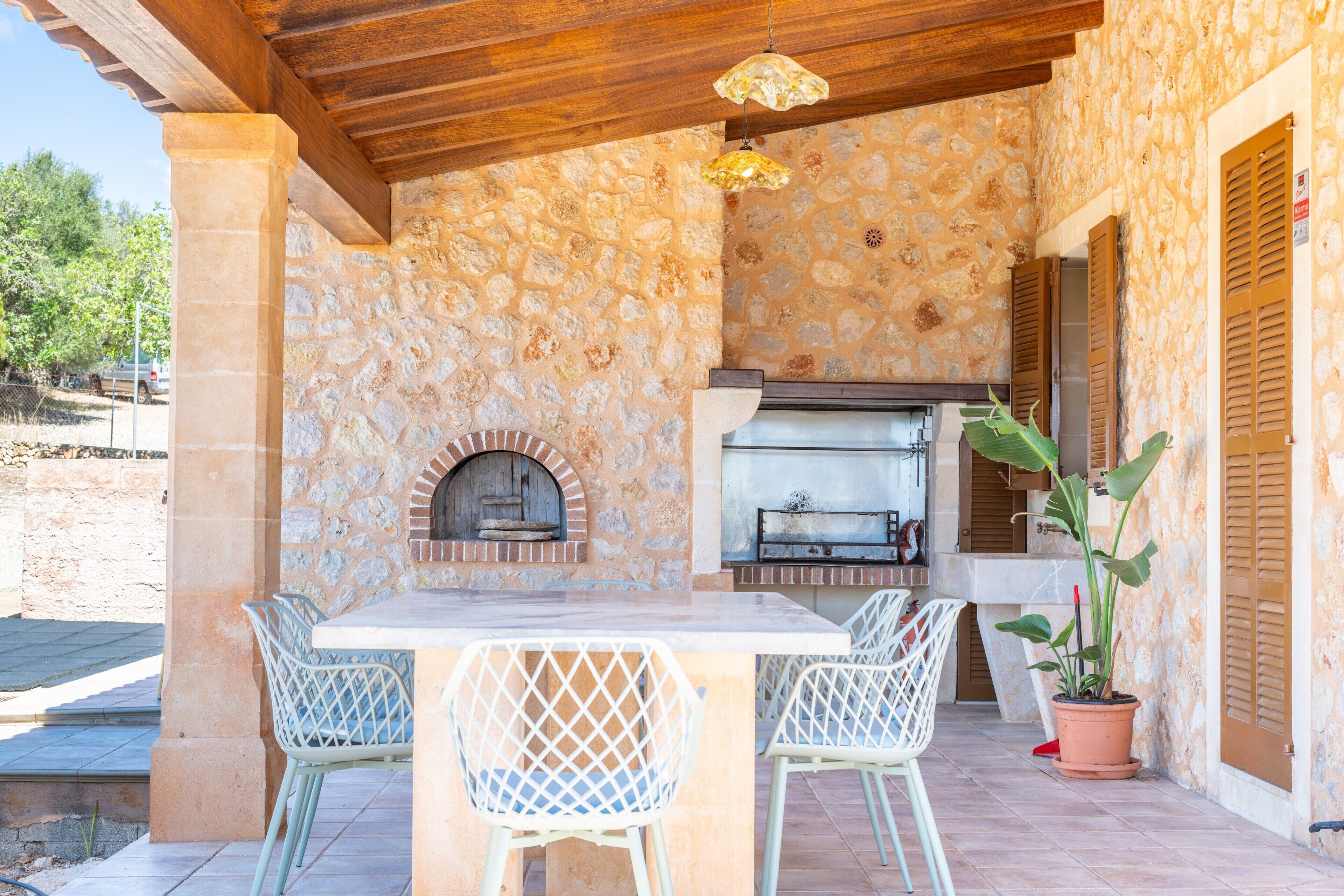 An outdoor kitchen with stone walls, a brick oven, and a grill. There are two white chairs and a table.