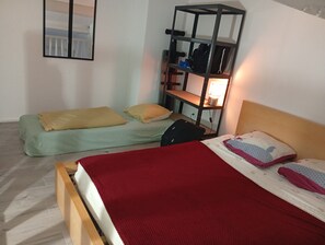 Ein Zimmer mit zwei Betten, eines mit roter Bettdecke und das andere mit gelber Decke. Ein Regal mit einer Lampe darauf.