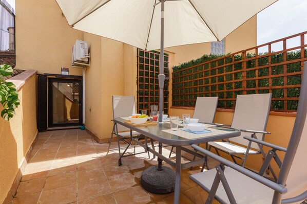 Un patio exterior con una mesa, sillas, un gran paraguas y un muro con un enrejado.