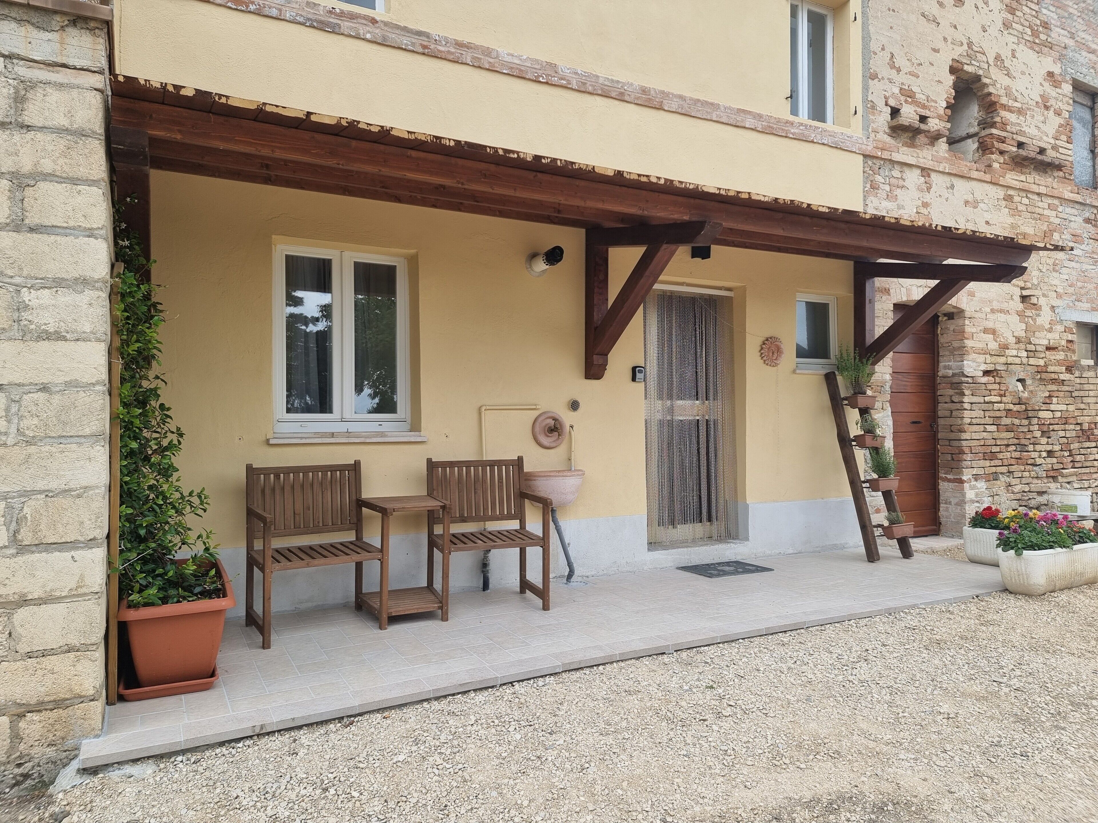 A building with a wooden pergola, two wooden chairs, a potted plant, and a door with a window.