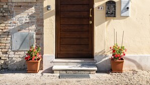 Une porte en bois avec une poignée en métal, de part et d’autre de laquelle se trouvent deux plantes en pot ornées de fleurs rouges, et une enseigne au-dessus de la porte indiquant « B&B Bianca Lana ».
