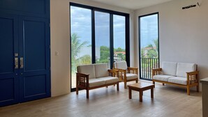 A spacious room with a large blue door, two wooden sofas with cushions, a small wooden table, and large windows offering a view of greenery and buildings.