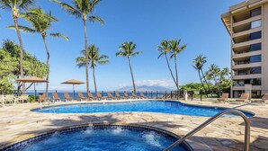 Une piscine entourée de chaises longues et de palmiers, elle-même bordée d’une terrasse en pierre et d’un bâtiment doté de balcons.
