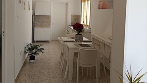 Een smalle gang die uitkomt op een keuken met witte kasten, een eettafel met stoelen en een raam met jaloezieën.