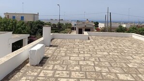 Een dak terras met betegelde vloer, witte muren en uitzicht op zee en andere gebouwen.