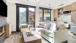 A modern living room with a sofa, coffee table, and a fireplace. There is a balcony with a view of buildings. The kitchen has modern appliances and a large window.