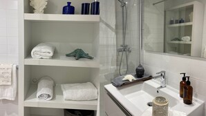 Ein modernes Badezimmer mit weißen Fliesen, einer Duschkabine aus Glas, einem weißen Waschbecken mit Spiegel sowie Regalen mit Handtüchern und Pflegeprodukten.
