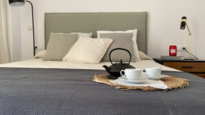 Ein ordentlich bezogenes Bett mit grauer Decke, zwei weiße Tassen, ein schwarzer Teekessel und eine rote Kaffeemaschine auf einem Nachttisch.