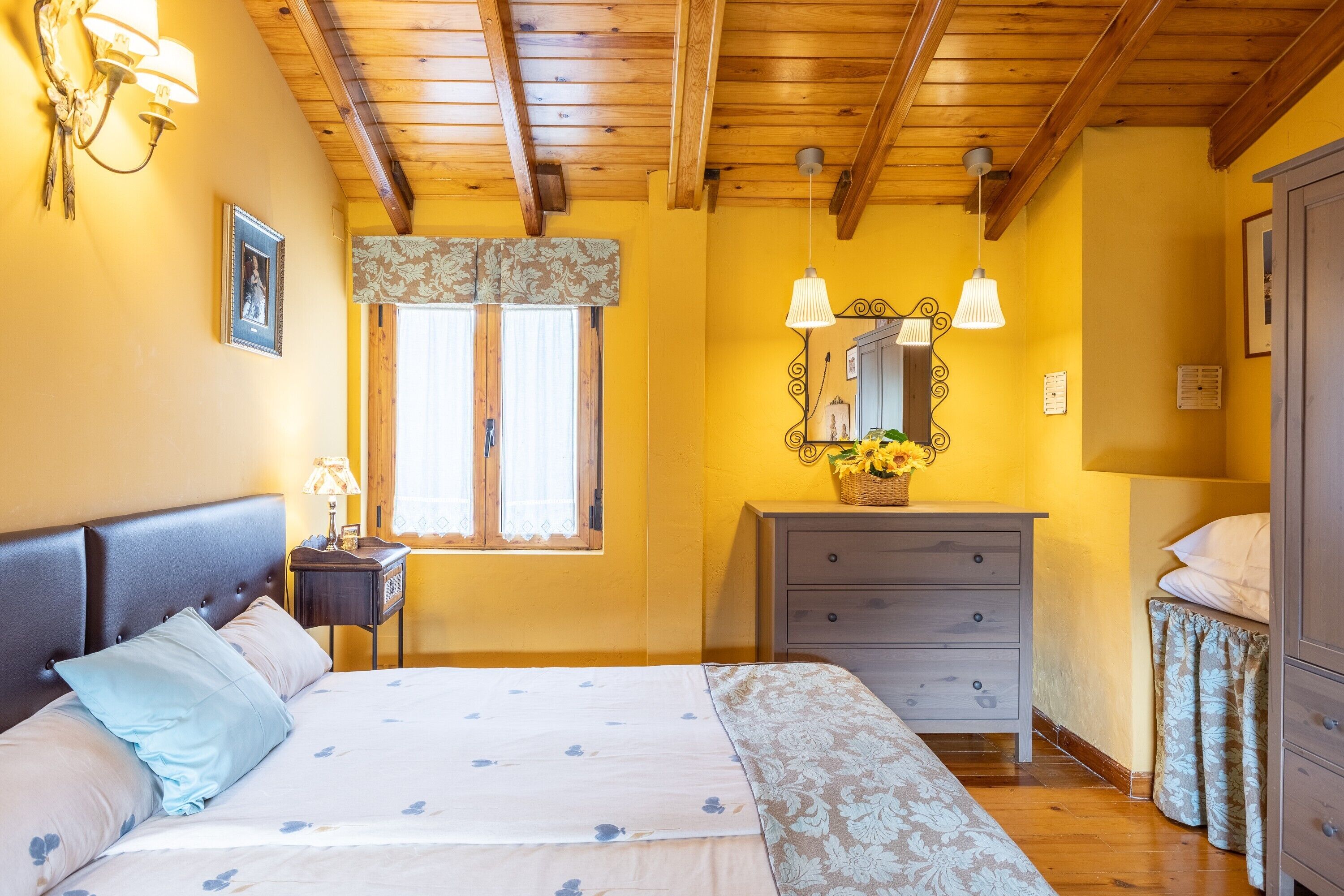 Een slaapkamer met een bed, bedlampje, dressoir, spiegel en raam met gordijnen.