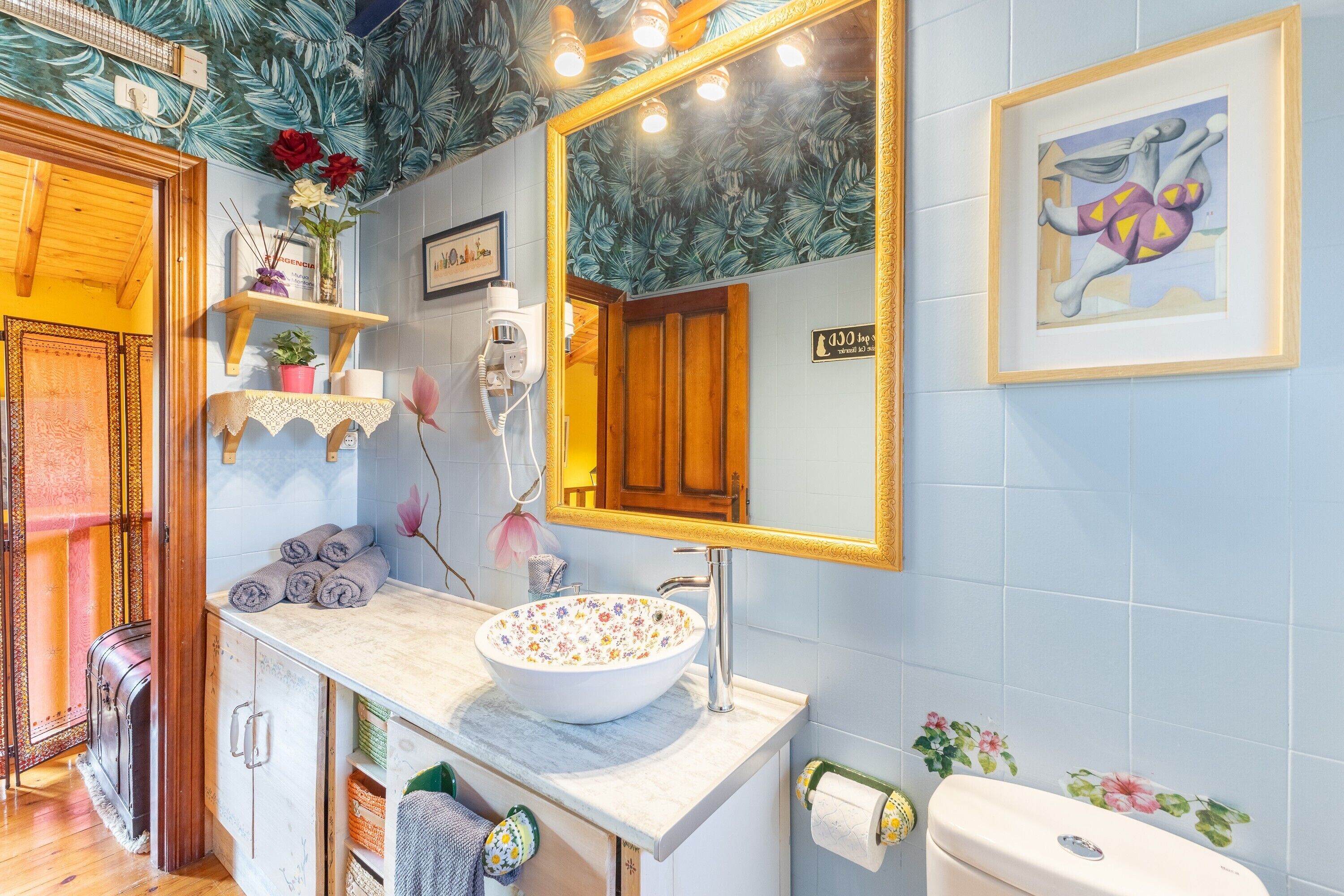 Un baño con lavamanos estampado con motivos florales, un armario de madera, un espejo con marco dorado y una obra de arte enmarcada en la pared.