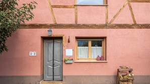 A traditional building with a wooden-framed window, a door with a number plaque, and a small flower arrangement on the ground.