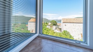 Une pièce dotée d’une grande fenêtre donnant sur un village côtier, avec ses bâtiments en pierre et un ciel d’un bleu éclatant.