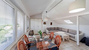 Un salon moderne avec une table à manger en verre, des chaises en osier, un canapé et une grande fenêtre avec des stores.