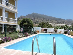 A swimming pool with a view of mountains, a building with balconies, and a fenced garden area.