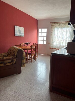 Una habitación con una mesa de comedor, sillas, un sofá, un ventanal con cortinas y una zona de cocina.