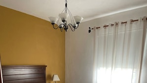 En dobbeltseng med to puder, et natbord med lampe, en lysekrone og et vindue med gardiner.
