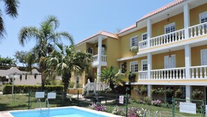 A swimming pool with a fenced area, surrounded by a two-story building with balconies, palm trees, and a clear sky. - Apartment for 5 with private terrace, shared pool & Wi-Fi in Los Llanos (Los Llanos)