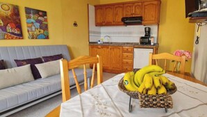 A compact kitchen with wooden cabinets, a dining table with a banana basket, and a sofa in the living area. - Apartment for 5 with private terrace, shared pool & Wi-Fi in Los Llanos (Los Llanos)