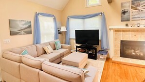 A living room with a sectional sofa, a flat-screen TV, a fireplace, and a window with curtains. - Charming Cape Cod summer house conveniently located  with plenty to offer. (Mashpee)
