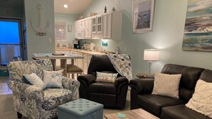 A living room with a sofa, two armchairs, a coffee table, and a small ottoman. There is a kitchen area in the background with white cabinets and a dining table. - New, update, waterfront condo. Relaxing water sounds and view. (Pensacola Beach)