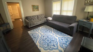A living room with a gray sofa, a patterned rug, a painting on the wall, and a door leading to another room.