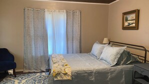 A bedroom with a bed, a chair, a desk, a window with curtains, and a painting on the wall.