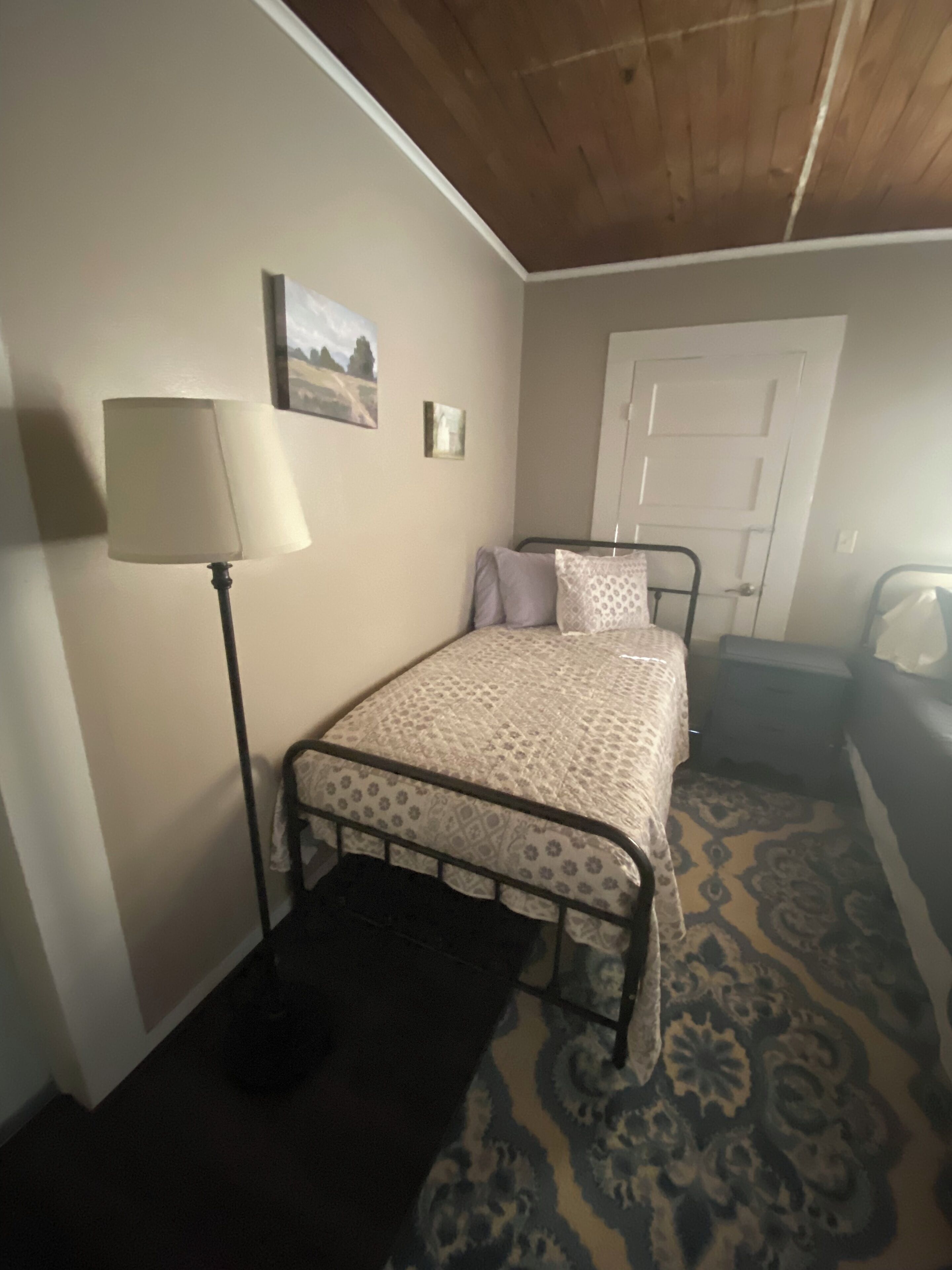 Un dormitorio con una cama, una mesita de noche, una lámpara, una puerta y una alfombra estampada.