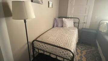 Un dormitorio con una cama, una mesita de noche, una lámpara, una puerta y una alfombra estampada.
