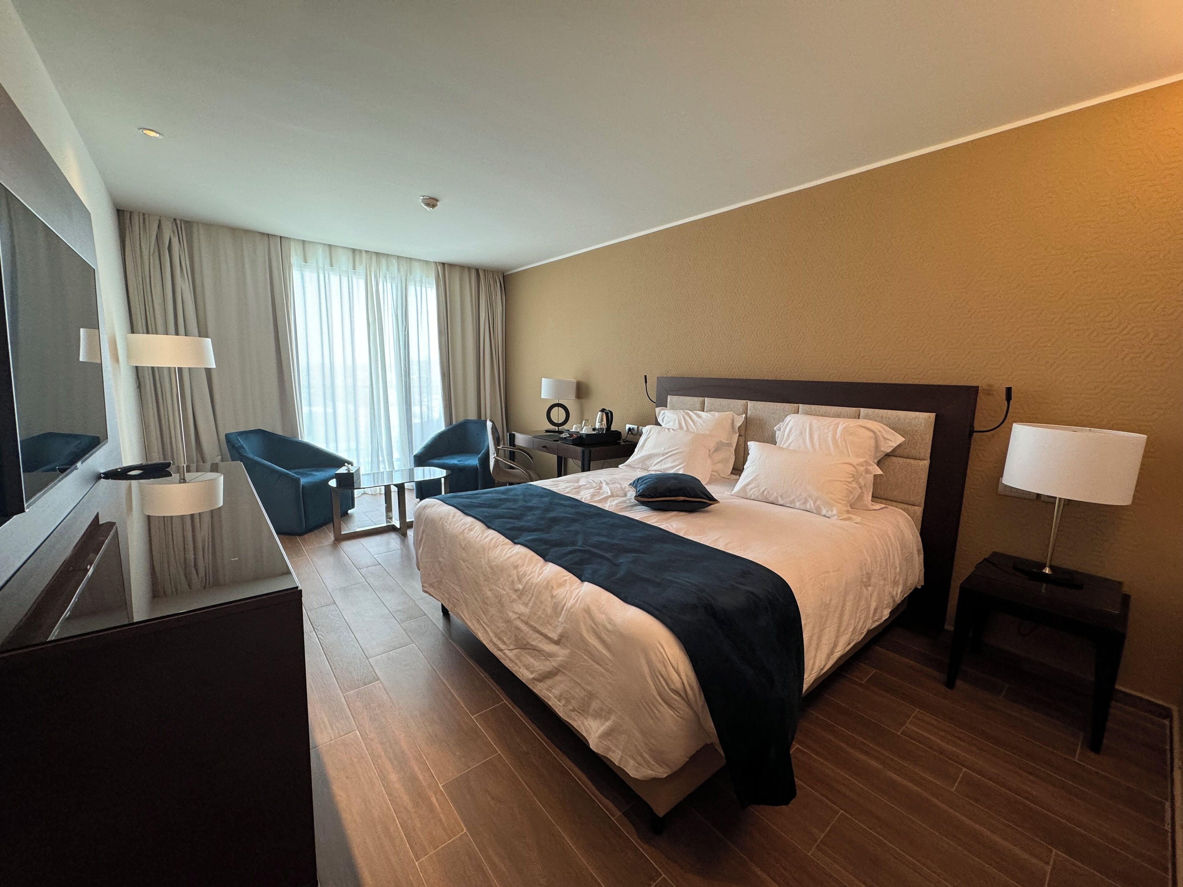 A hotel room with a bed, two chairs, a desk, a TV, and a lamp.