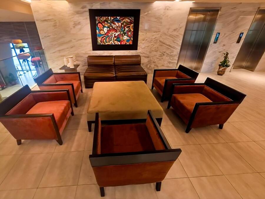 A lobby with a central seating area featuring a long table and multiple cushioned chairs, a large abstract painting on the wall, and an elevator.