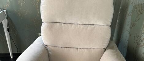 Un fauteuil inclinable de couleur beige, doté d’un appareil de massage, placé contre un mur recouvert d’un papier peint à motifs floraux.