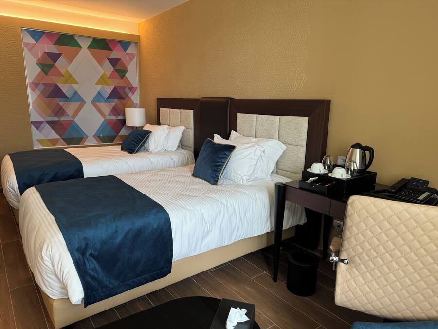 Quarto de hotel com duas camas, uma mesa com telefone, uma cadeira, um cesto de lixo e uma parede com arte decorativa de padrão geométrico.